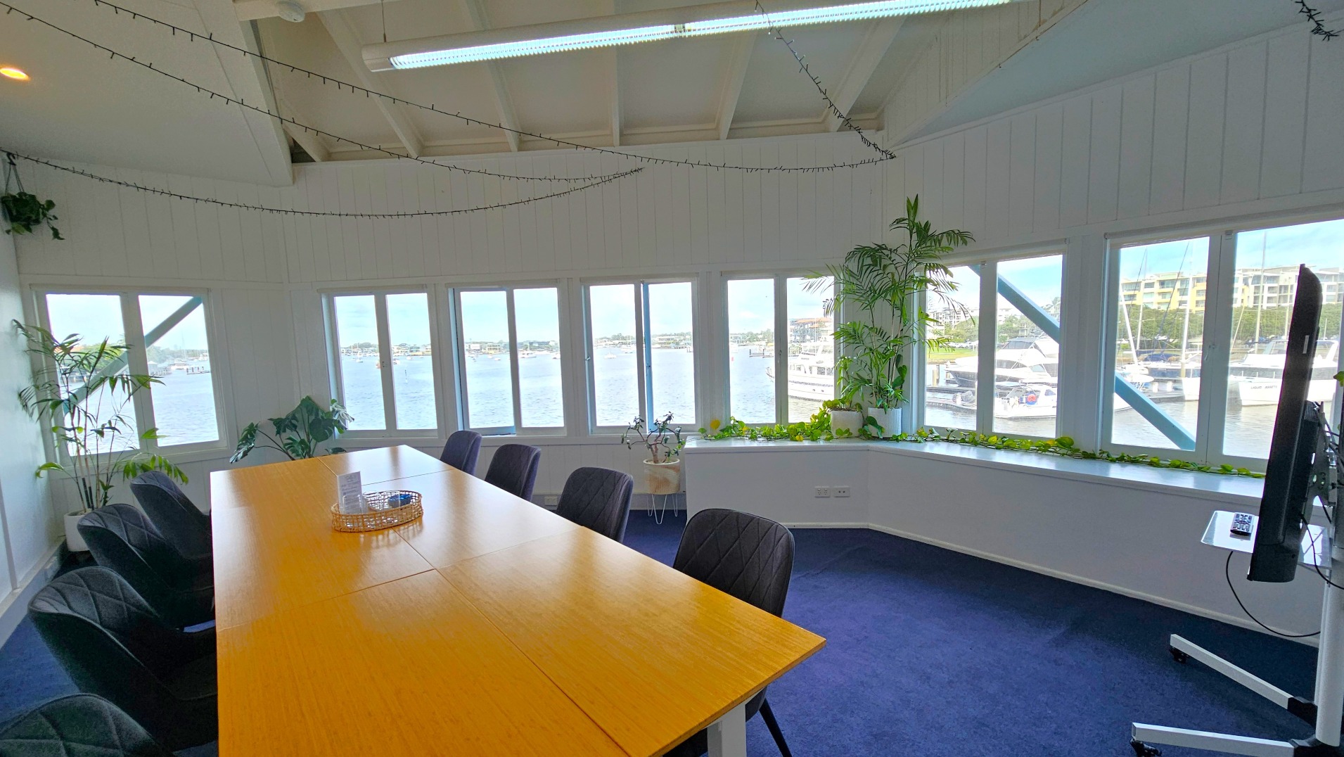 A conference room with a long wooden table surrounded by black chairs on a blue carpet. Large windows line the walls, offering views of a marina with boats and buildings. Potted plants are placed along the window sills and corners, adding greenery. The ceiling is high with exposed beams and string lights. A flat-screen monitor is mounted on a stand to the right.