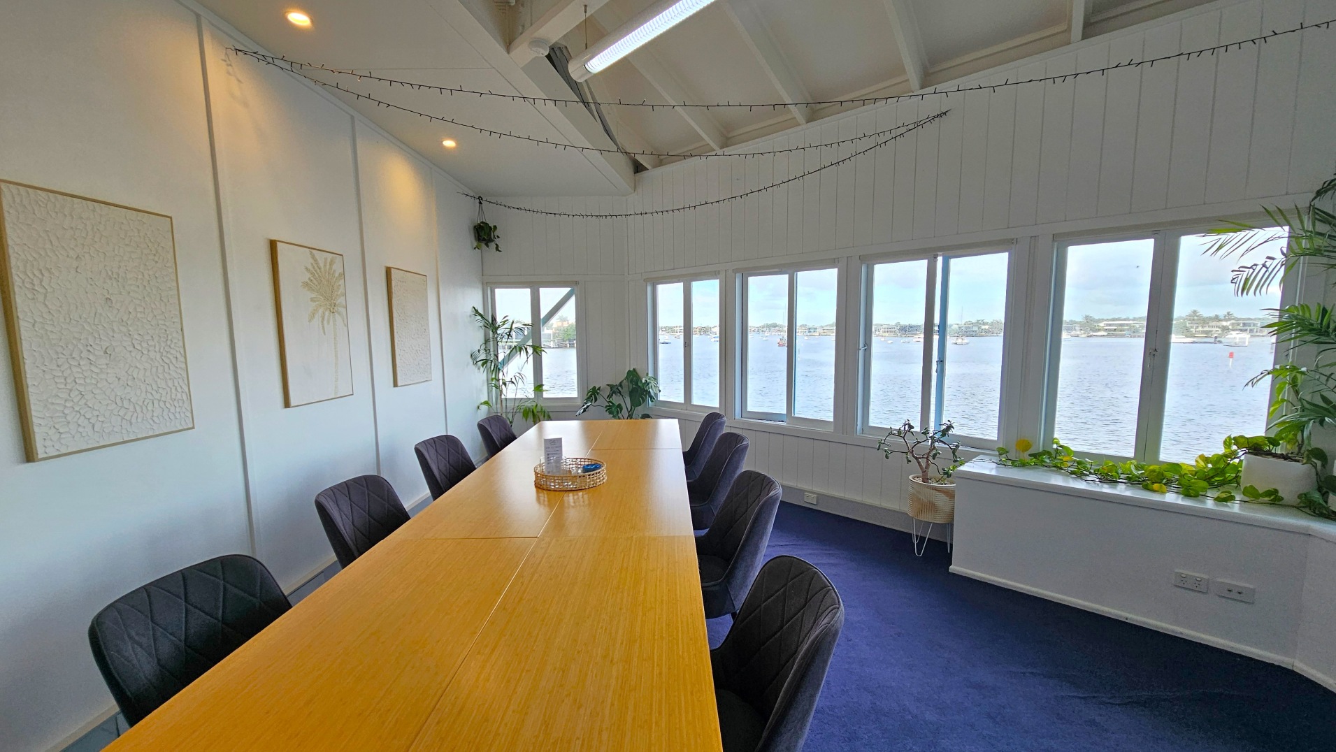 A meeting room with a long wooden table surrounded by dark cushioned chairs. The room features large windows offering a view of a body of water and distant buildings. The walls are white with three framed artworks. Potted plants are placed near the windows and along the ledge. The ceiling is high with exposed beams and string lights. The floor is carpeted in dark blue.