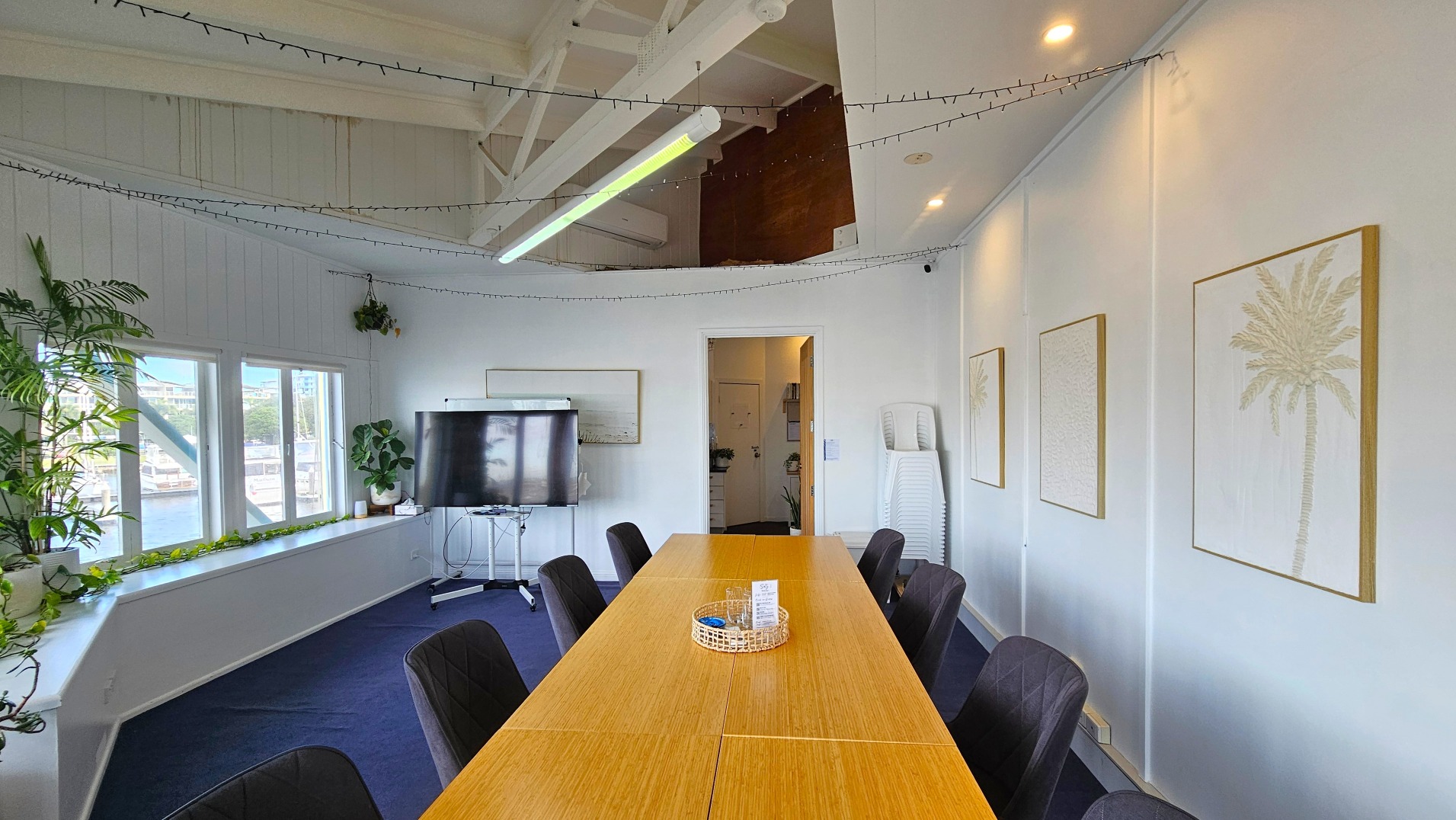 A meeting room with a long wooden table surrounded by black chairs. The room features white walls adorned with framed botanical prints. A large flat-screen TV on a stand is positioned near a doorway. Windows along one wall allow natural light, with potted plants on the windowsill. The ceiling has exposed beams and string lights. A stack of white chairs is in the corner, and the floor is carpeted in dark blue.