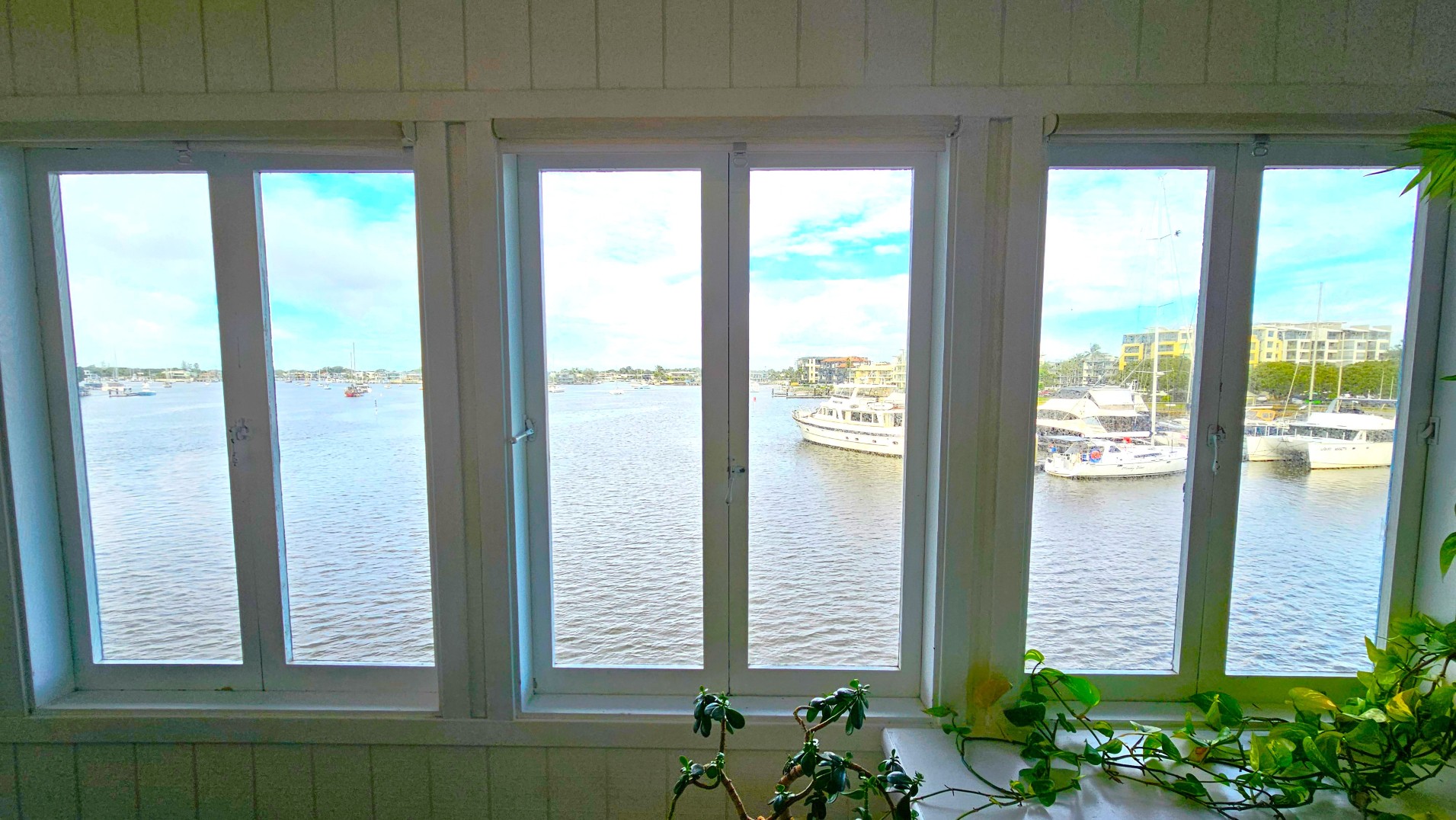 A view through four large vertical windows showing a marina with several boats docked on calm water. The windows are framed in white, and a plant with green leaves is visible on the windowsill. Beyond the marina, there are low-rise buildings and a few scattered trees. The interior wall is clad in white panelling, adding a clean, nautical feel to the space.