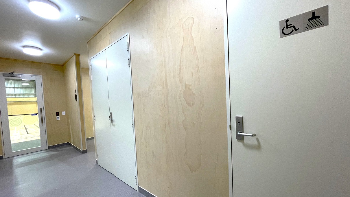 Accessible bathroom and shower at the Gallagher Recreation Centre. The accessible bathroom has a disabled symbol and a shower head with water droplets on the off-white painted door. Before the accessible bathroom is a off-white painted double door and a hallway right after the glass entrance doors. All doors have a lever type, stainless steel door handle. Accessible bathroom and shower at the Gallagher Recreation Centre. The accessible bathroom has a disabled symbol and a shower head with water droplets on the off-white painted door. Before the accessible bathroom is a off-white painted double door and a hallway right after the glass entrance doors. All doors have a lever type, stainless steel door handle.