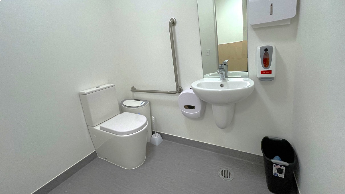 The bathroom has a grey floor and a white wall with dark brown painted bottom. On the left side of the toilet is a white/grey air purifier, a white toilet brush, and grab bars fixed to the white wall. There is also a low-set hand basin, a mirror, a liquid hand soap dispenser, and a toilet paper roll dispenser. A small black garbage bin is placed against the wall opposite the toilet. The bathroom has a grey floor and a white wall with dark brown painted bottom. On the left side of the toilet is a white/grey air purifier, a white toilet brush, and grab bars fixed to the white wall. There is also a low-set hand basin, a mirror, a liquid hand soap dispenser, and a toilet paper roll dispenser. A small black garbage bin is placed against the wall opposite the toilet.