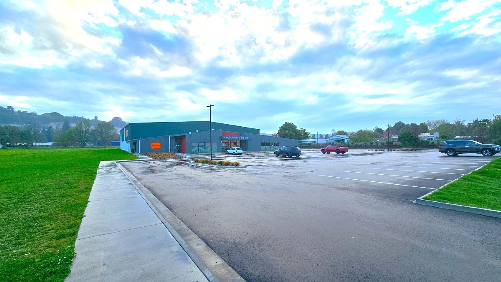 A large parking area with a few cars scattered around. On the left, there's a wide expanse of green grass. In the centre, a single-storey building with a dark green exterior and a red sign is visible. The foreground features a wet pavement, suggesting recent rain. The background includes trees and distant hills under a cloudy sky. A large parking area with a few cars scattered around. On the left, there's a wide expanse of green grass. In the centre, a single-storey building with a dark green exterior and a red sign is visible. The foreground features a wet pavement, suggesting recent rain. The background includes trees and distant hills under a cloudy sky.