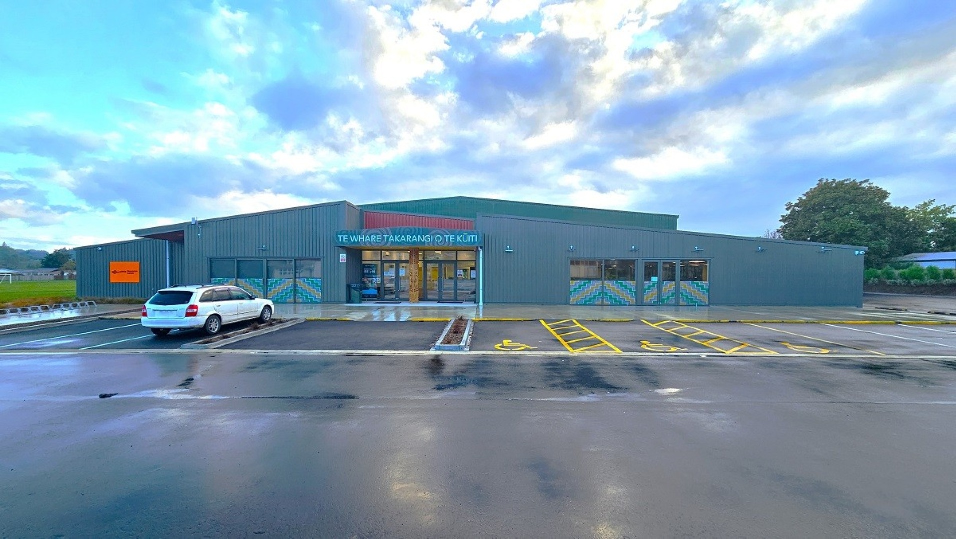 A modern, single-storey building with a grey exterior and a large entrance in the centre. The entrance has glass doors and a sign above it. The building is surrounded by a wet car park with marked parking spaces, including a white car parked on the left. The sky is overcast, and there are trees visible in the background on the right. The ground is wet, suggesting recent rain. A modern, single-storey building with a grey exterior and a large entrance in the centre. The entrance has glass doors and a sign above it. The building is surrounded by a wet car park with marked parking spaces, including a white car parked on the left. The sky is overcast, and there are trees visible in the background on the right. The ground is wet, suggesting recent rain.