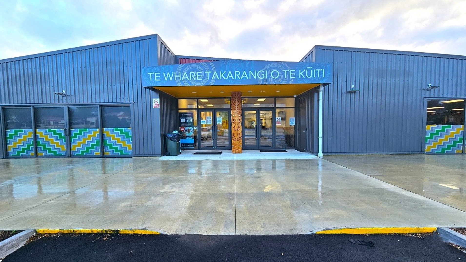 A modern building with a grey exterior and a blue sign above the entrance reading "Te Whare Takarangi o Te Kuiti". The entrance features large glass doors and a wooden carved pillar. On either side of the entrance, there are windows with colourful geometric patterns. The ground is wet, suggesting recent rain, and the pavement is smooth and reflective. The roof is flat, and the overall structure is symmetrical. A modern building with a grey exterior and a blue sign above the entrance reading "Te Whare Takarangi o Te Kuiti". The entrance features large glass doors and a wooden carved pillar. On either side of the entrance, there are windows with colourful geometric patterns. The ground is wet, suggesting recent rain, and the pavement is smooth and reflective. The roof is flat, and the overall structure is symmetrical.