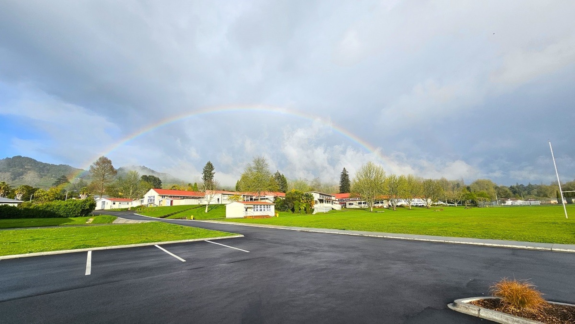A paved parking area in the foreground with white lines marking spaces. To the right, a small patch of grass with a few plants. In the background, a series of white buildings with red roofs are surrounded by green lawns and trees. A faint rainbow arcs across the sky above the buildings. Hills and more trees are visible in the distance. A paved parking area in the foreground with white lines marking spaces. To the right, a small patch of grass with a few plants. In the background, a series of white buildings with red roofs are surrounded by green lawns and trees. A faint rainbow arcs across the sky above the buildings. Hills and more trees are visible in the distance.
