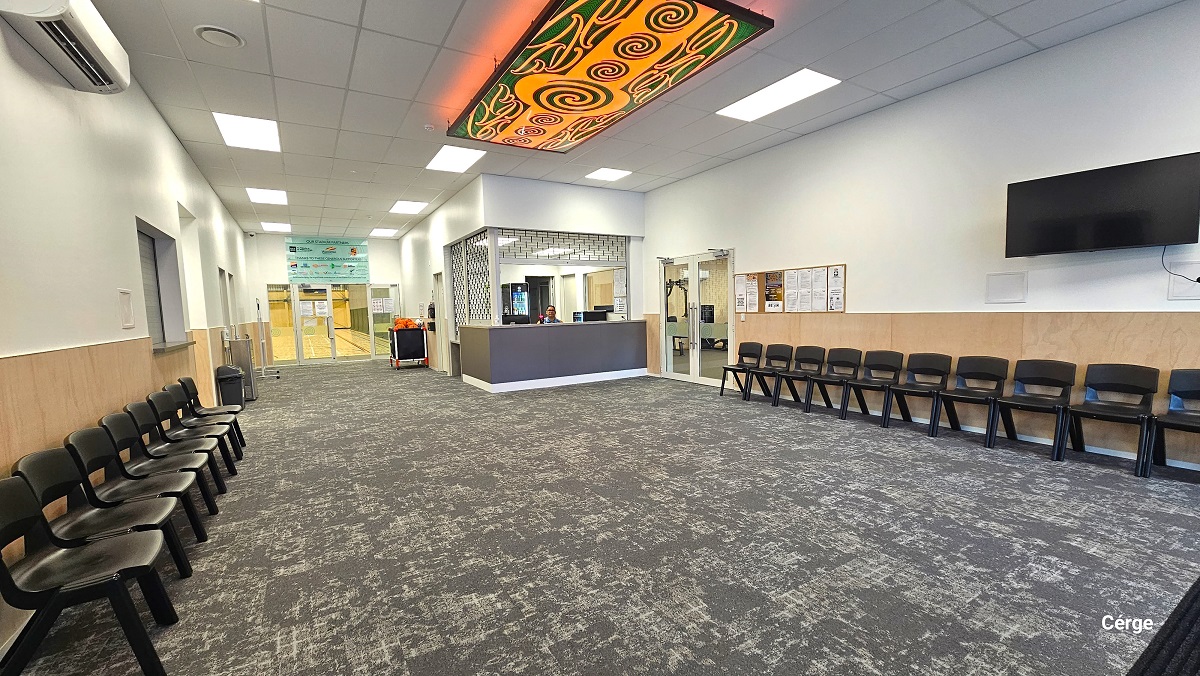 Grey carpet tiles flooring in the reception area. Black plastic chairs are placed against the brown-painted part of the wall on either side of the entrance. The ceiling is painted white with alternating rectangular lighting patterns and a green and yellow-orange Maori artwork. A TV is mounted on the right wall, and further beyond is a wooden bulletin board. To the right of the reception desk is the gym, to the left is the kitchen and bathrooms, while directly ahead is the stadium. Grey carpet tiles flooring in the reception area. Black plastic chairs are placed against the brown-painted part of the wall on either side of the entrance. The ceiling is painted white with alternating rectangular lighting patterns and a green and yellow-orange Maori artwork. A TV is mounted on the right wall, and further beyond is a wooden bulletin board. To the right of the reception desk is the gym, to the left is the kitchen and bathrooms, while directly ahead is the stadium.