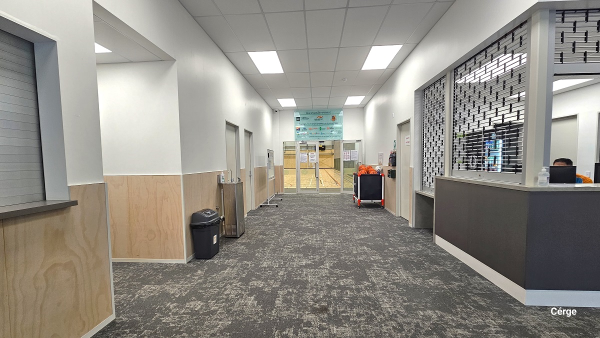 The reception desk at Gallagher Recreation Centre can be distinguished by the aluminum roller grille and a marble countertop with a grey-painted wall. There is another door next to the reception desk, while there are multiple doors opposite it. A small black garbage bin, a water fountain, and a mobile whiteboard can be found on the left side of the hallway leading to the stadium. On the right is a black storage ball rack. There is a light blue glass signboard atop the glass doors of the stadium. The reception desk at Gallagher Recreation Centre can be distinguished by the aluminum roller grille and a marble countertop with a grey-painted wall. There is another door next to the reception desk, while there are multiple doors opposite it. A small black garbage bin, a water fountain, and a mobile whiteboard can be found on the left side of the hallway leading to the stadium. On the right is a black storage ball rack. There is a light blue glass signboard atop the glass doors of the stadium.