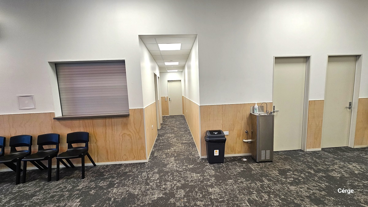 Kitchen and bathroom section at Gallagher Recreation Centre. On the left side, before the hallway, is a small window with an extended flat surface and several black plastic chairs. On the right side is the small black garbage bin, a water fountain, and two off-white painted doors. The image also shows a grey carpet tiles flooring and a combination of a brown wooden lower part and a white-painted upper wall and ceiling. Kitchen and bathroom section at Gallagher Recreation Centre. On the left side, before the hallway, is a small window with an extended flat surface and several black plastic chairs. On the right side is the small black garbage bin, a water fountain, and two off-white painted doors. The image also shows a grey carpet tiles flooring and a combination of a brown wooden lower part and a white-painted upper wall and ceiling.