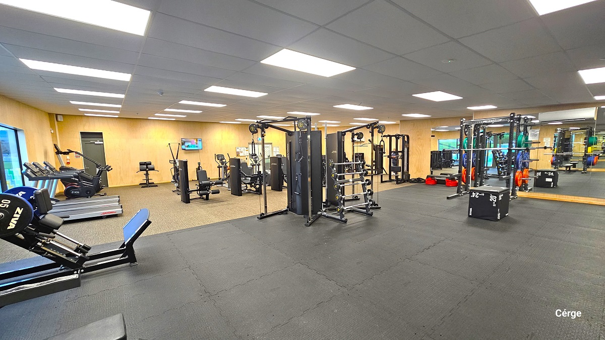 A closer look at the space inside the gymnasium near the TV mounted on a wall. The treadmills are aligned next to each other on the wall near the exit glass doors. This image also shows the large floor-to-ceiling mirror and an air conditioning unit inside the gymnasium on the right wall standing opposite the entrance doors. There is a large black box in the middle of the black gym mat floor. A closer look at the space inside the gymnasium near the TV mounted on a wall. The treadmills are aligned next to each other on the wall near the exit glass doors. This image also shows the large floor-to-ceiling mirror and an air conditioning unit inside the gymnasium on the right wall standing opposite the entrance doors. There is a large black box in the middle of the black gym mat floor.