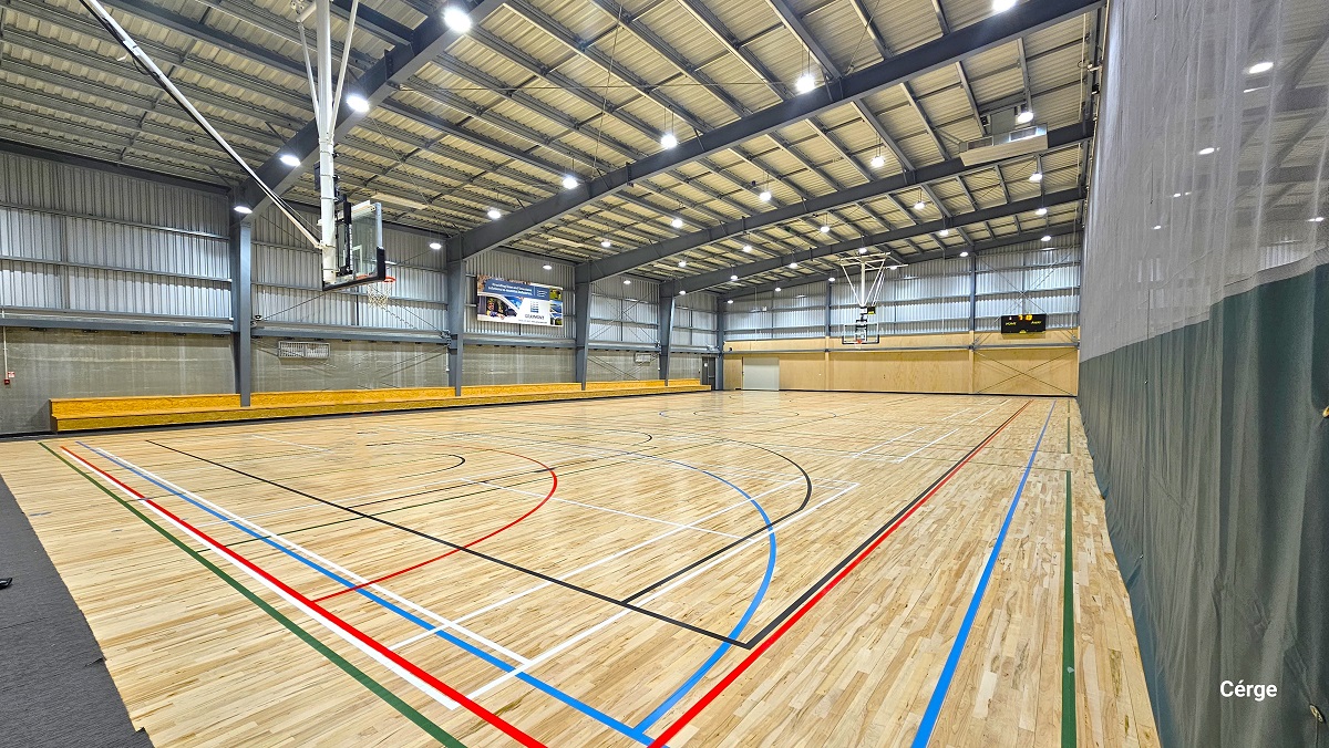 Side view of one of the basketball courts in the Sports stadium. The other court is separated by a curtain with a dark lower part. The floor surface changed from carpet tiles to timber floors with a highly polished finish which creates a low-level glare from the fluorescent lighting. The stadium has a very high ceiling made of off-white corrugated iron and wide grey steel beams. There is a two-tiered seating along two sides of the stadium. Side view of one of the basketball courts in the Sports stadium. The other court is separated by a curtain with a dark lower part. The floor surface changed from carpet tiles to timber floors with a highly polished finish which creates a low-level glare from the fluorescent lighting. The stadium has a very high ceiling made of off-white corrugated iron and wide grey steel beams. There is a two-tiered seating along two sides of the stadium.