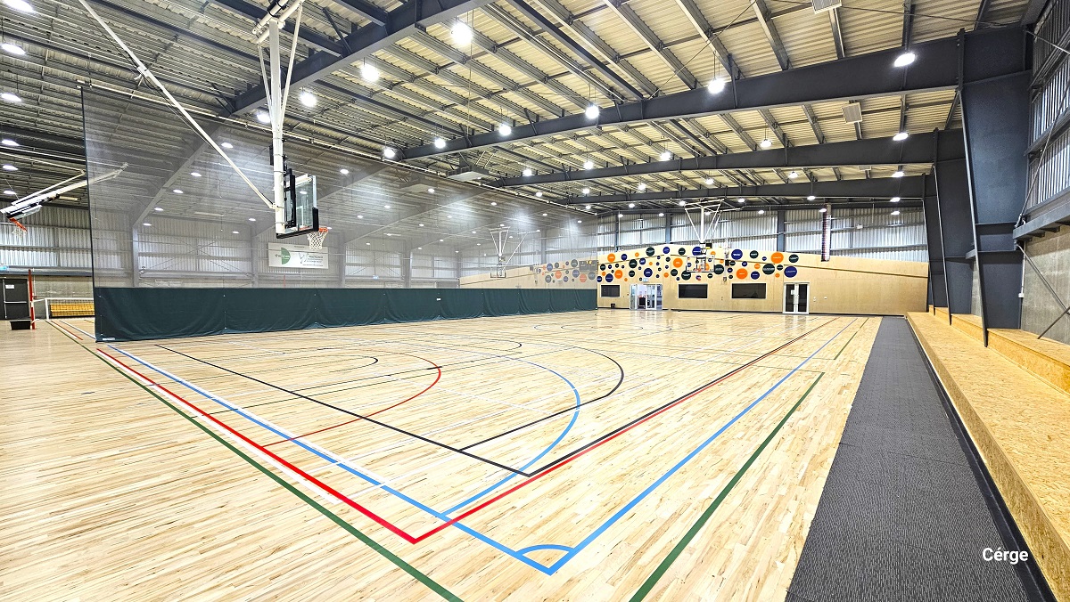 View of the basketball court standing in front of the two-tiered brown seating on the right side. There is a grey mat that separates the court from the seating area. Two glass doors at the back with white metal frames can be found within the brown-painted wall at the back with colorful circular designs. The other court has a low net wrapped around the red pole at the opposite ends. View of the basketball court standing in front of the two-tiered brown seating on the right side. There is a grey mat that separates the court from the seating area. Two glass doors at the back with white metal frames can be found within the brown-painted wall at the back with colorful circular designs. The other court has a low net wrapped around the red pole at the opposite ends.