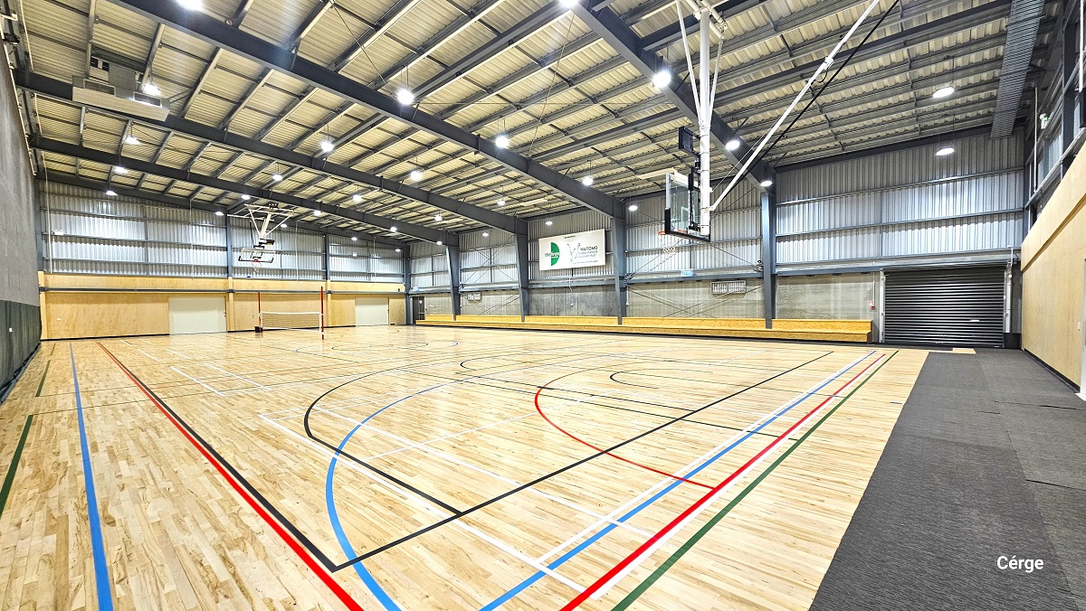 View of the second court with a low white/black wrapped around two red poles at opposite sides. The right end of the court is covered with grey mats, while the rest of the court has polished timber floors with red, black, blue, white, and green lines. There is also a two-tiered brown seating along the stadium side. On the upper part of the grey corrugated wall to the right is a large, white advertisement poster. View of the second court with a low white/black wrapped around two red poles at opposite sides. The right end of the court is covered with grey mats, while the rest of the court has polished timber floors with red, black, blue, white, and green lines. There is also a two-tiered brown seating along the stadium side. On the upper part of the grey corrugated wall to the right is a large, white advertisement poster.