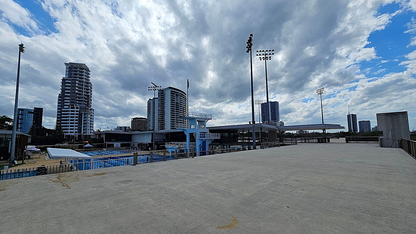 Wide Open Space and Possible Quiet Area and skyline.  Level 1 Tiered Seating.  Gold Coast Aquatic Centre.  A large, open concrete area with rows of empty seats along the left side. Tall floodlights are visible in the background, along with some trees. On the right, there is a covered structure with a roof supported by metal beams. A railing runs along the right edge of the area. The sky is clear with a few clouds.