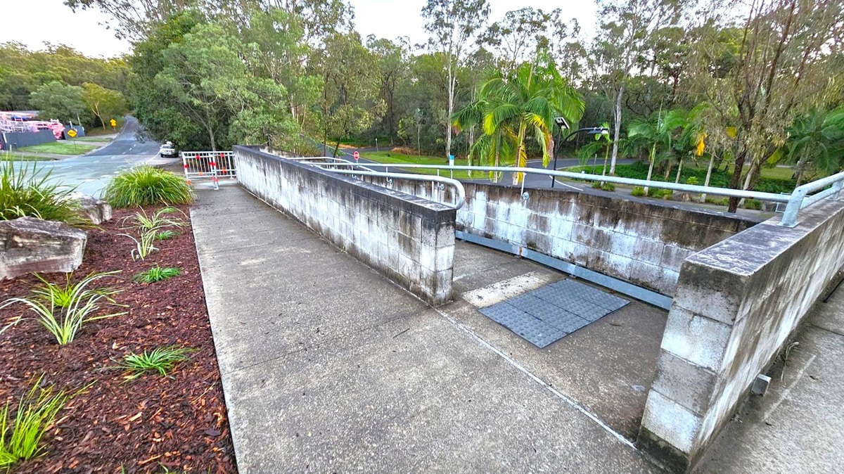 A concrete pathway with a metal railing on the right leads downwards, bordered by a low, weathered brick wall. On the left, there is a landscaped area with small plants and mulch. In the background, a road curves to the left, surrounded by dense trees and greenery, including palm trees. A metal barrier is visible at the end of the path.