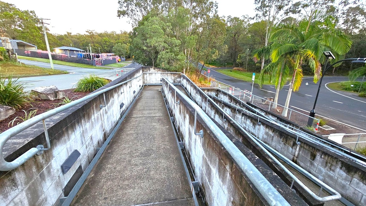 Concrete ramp with metal railings leading downwards, flanked by walls on both sides. On the left, a grassy area with rocks and a road leading uphill, bordered by a fence. On the right, palm trees and a curved road with a streetlamp. In the background, dense trees and a few buildings are visible. The area appears to be a suburban setting with a mix of natural and urban elements.