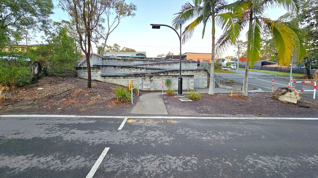 A paved path leads from the foreground across a road, flanked by low metal barriers and small plants. In the middle, a concrete ramp with railings ascends, with a person walking up. On the right, a palm tree stands near a fenced area with a large rock. In the background, trees and industrial buildings are visible, along with a parked vehicle. A streetlight with a solar panel is positioned near the ramp.