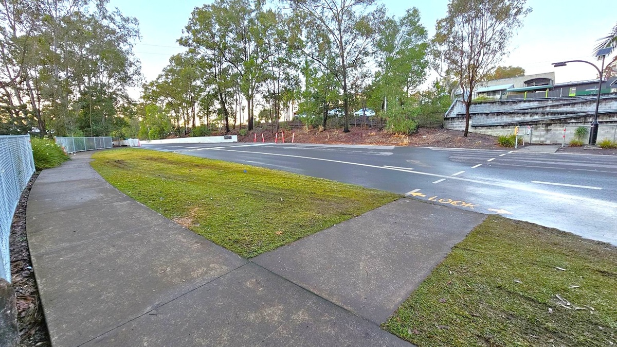 A curved concrete footpath runs along the left side, bordered by a chain-link fence and greenery. In the centre, a grassy area separates the path from a road. The road has a pedestrian crossing marked with "LOOK" in yellow. On the right, a train is visible on elevated tracks, supported by a concrete wall. Tall trees line the background, providing a natural backdrop.