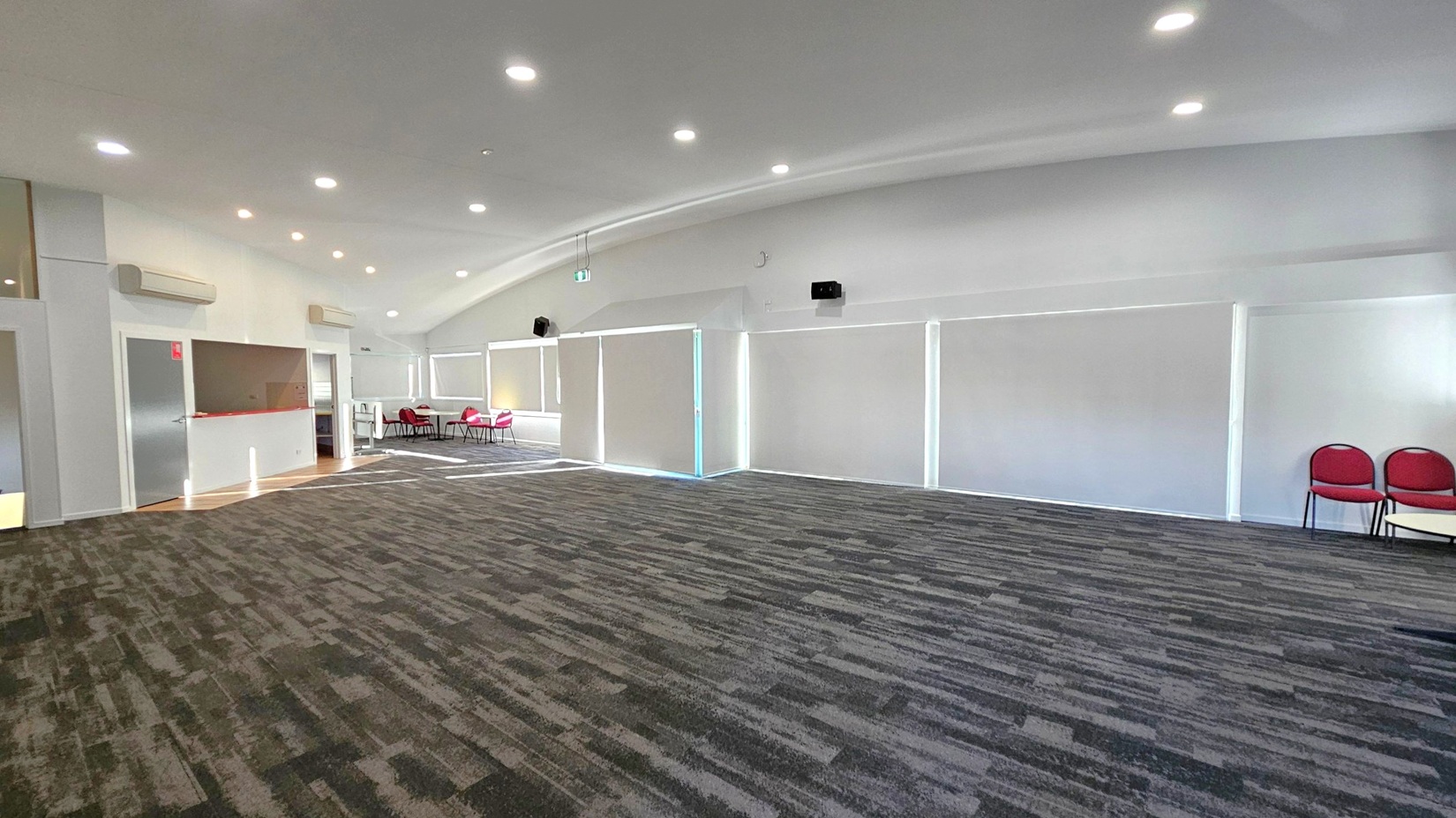 A spacious room with a patterned carpeted floor. On the left, there is a small counter area with a grey door and air conditioning units above. In the middle, large windows are covered with white blinds. On the right, two red chairs are placed against the wall. The ceiling is curved and features recessed lighting. In the background, a few more red chairs and tables are visible near another set of windows.