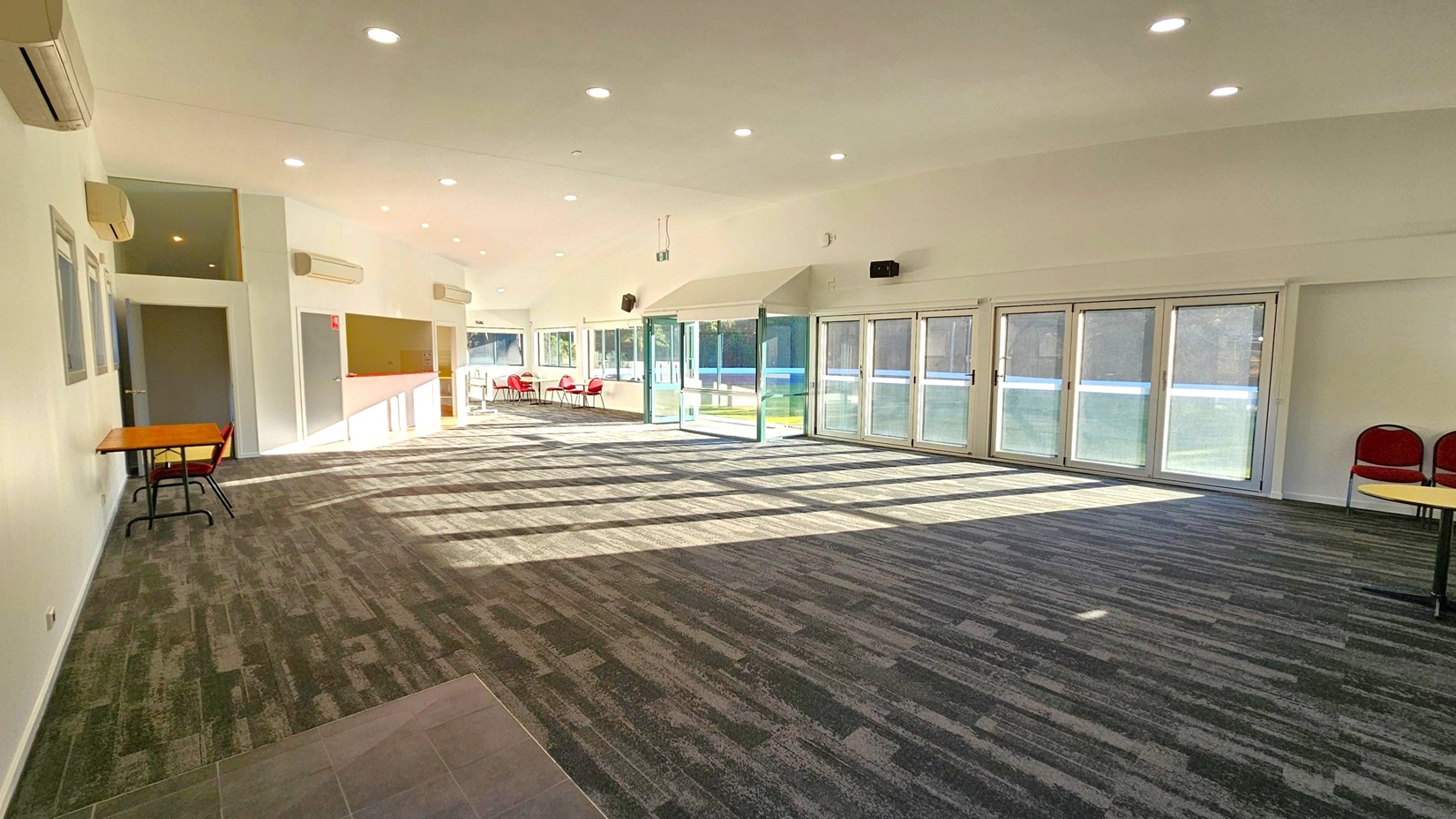 A spacious room with a patterned carpeted floor and a white ceiling with recessed lights. On the left, there are several air conditioning units mounted on the wall, a small wooden table with a chair, and a doorway. The right side features large glass doors with blue-tinted panels, allowing natural light to stream in. In the background, there are red chairs and tables near the glass doors, and a small counter area.