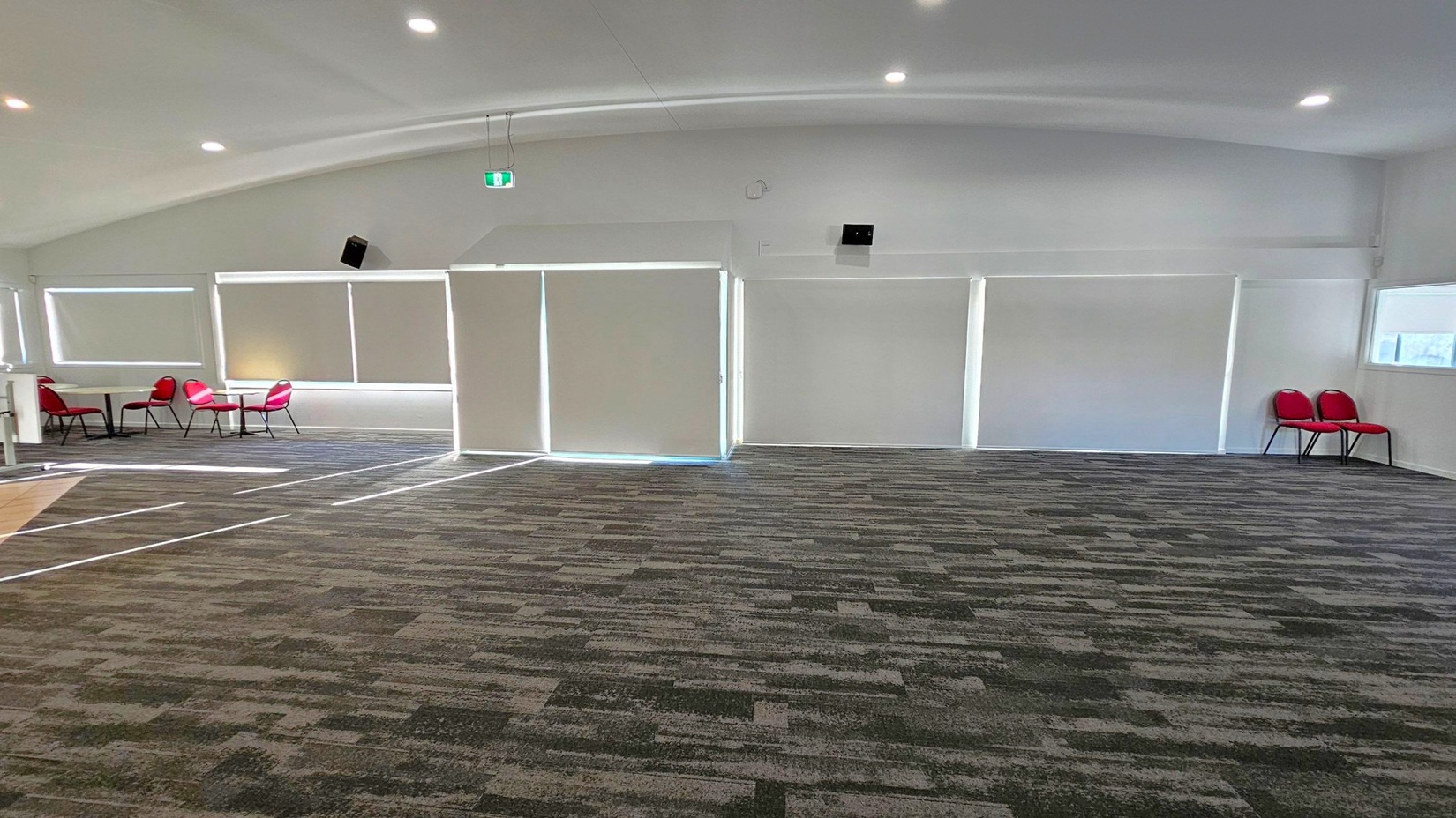 A spacious room with a patterned carpet floor and a white ceiling with recessed lighting. On the left, there are several red chairs around small tables near large windows with blinds. In the centre, the wall has large, closed blinds. On the right, two red chairs are placed against the wall near another window. The room has a modern and minimalistic design.