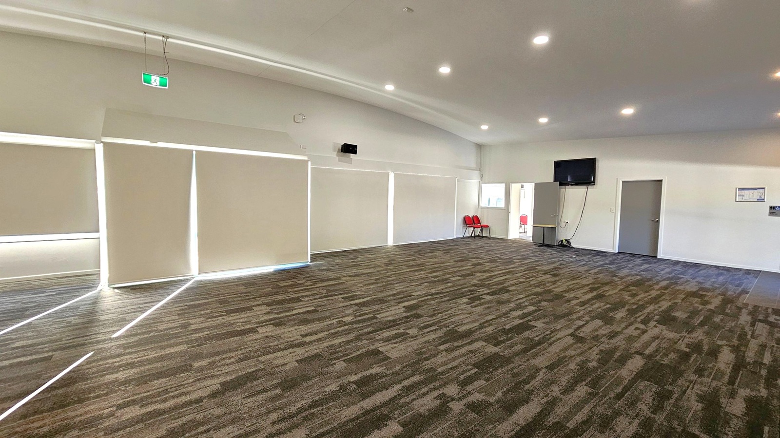 A spacious room with a patterned carpet in shades of brown and green. On the left, large windows are covered with blinds, allowing some light to filter through. The right side features a wall with a mounted television and two closed doors. Near the doors, a few red chairs are stacked. The ceiling is equipped with recessed lighting, and an emergency exit sign is visible above the windows. The room has a clean, modern appearance.