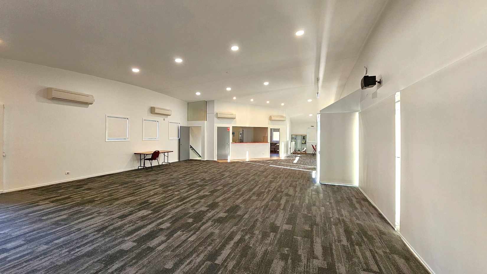 A spacious room with a patterned carpet in shades of green and brown. The ceiling is white with recessed lighting. On the left wall, there are three air conditioning units above small windows, and a table with a chair. The back wall features a counter with an open space behind it, leading to another room. The right wall has a mounted speaker and large windows with blinds. The room is well-lit and has a modern, minimalist design.