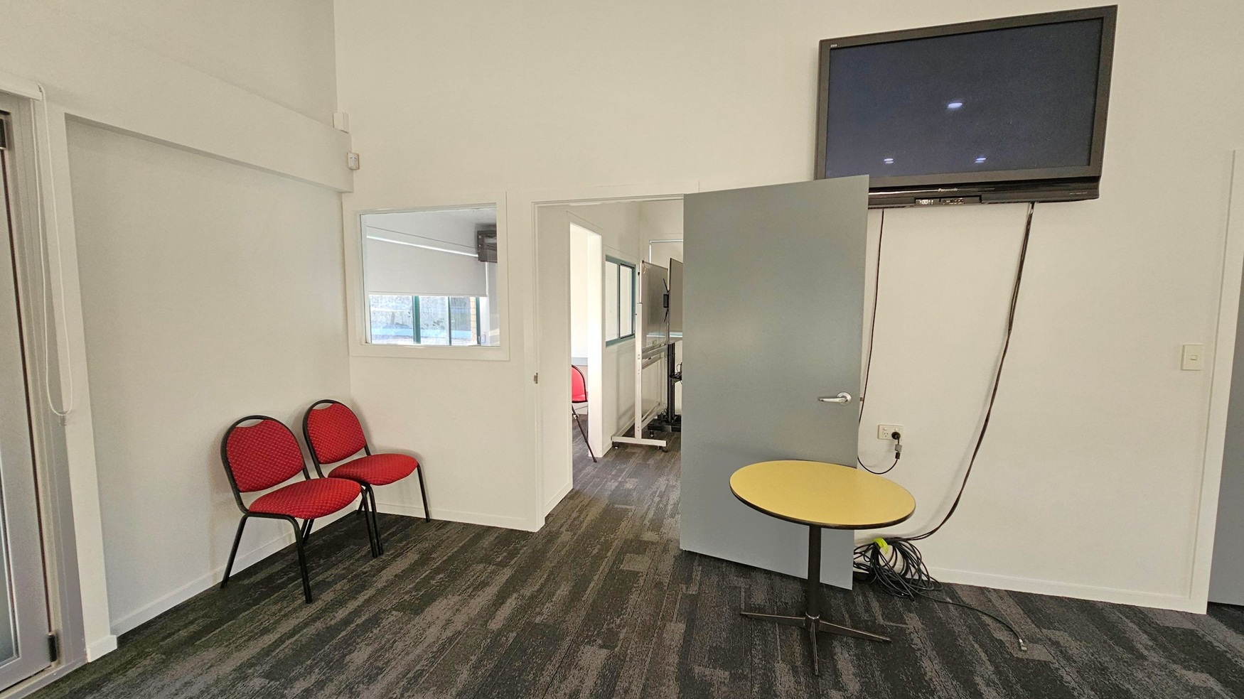 A room with a green and grey patterned carpet. On the left, two red chairs are placed against a white wall. In the centre, a door is partially open, leading to another room with more red chairs and a large screen. On the right, a wall-mounted television is above a small round table. Cables are visible near the floor. A window with a blind is partially visible through an interior window. The ceiling is white with recessed lights.