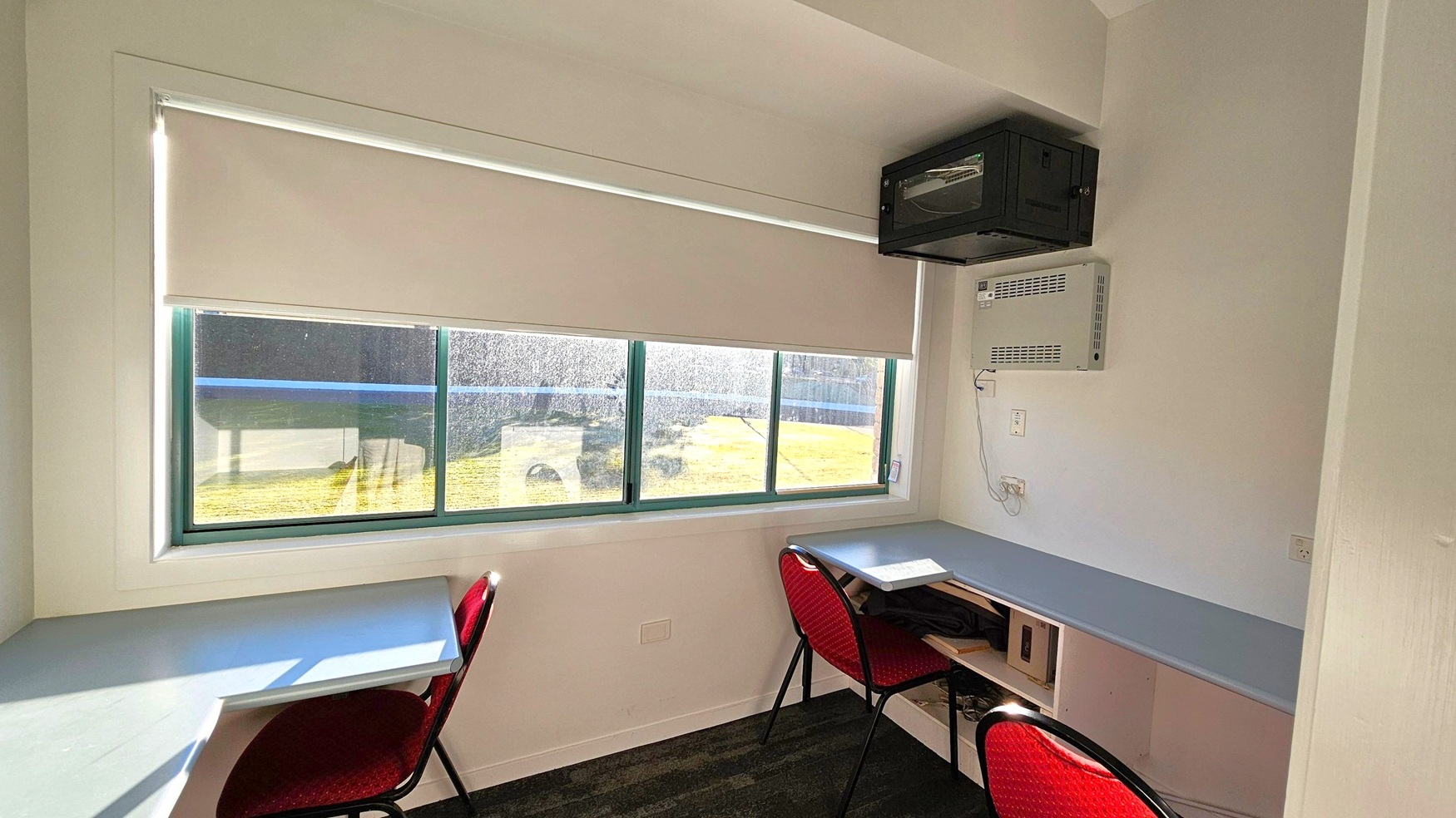 A small room with two desks and red chairs positioned against a wall with a large window. The window has a roller blind partially drawn, allowing some light to enter. Above the right desk, there is a black wall-mounted cabinet and an air conditioning unit. The floor is dark, and the walls are light-coloured. A power outlet and a light switch are visible on the wall near the air conditioning unit.