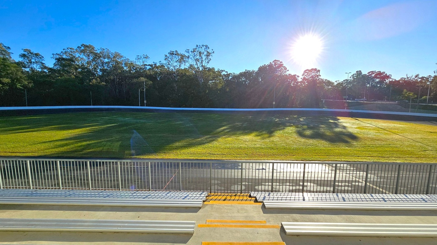 Bleachers in the foreground with metal railings and steps leading down to a grassy field. The field is surrounded by a white barrier, and beyond it, there are tall trees casting long shadows. The sun is low in the sky, creating a bright glare and illuminating the scene. The sky is clear and blue.