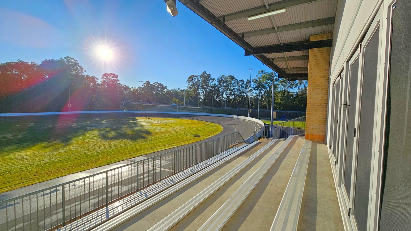 A sunlit outdoor sports track with a curved path is visible in the background, surrounded by trees. In the foreground, there are metal bleachers with a railing, positioned under a covered area with a corrugated metal ceiling. The right side features a brick wall and large windows or doors with mesh screens. The sun casts long shadows across the grassy area and track.