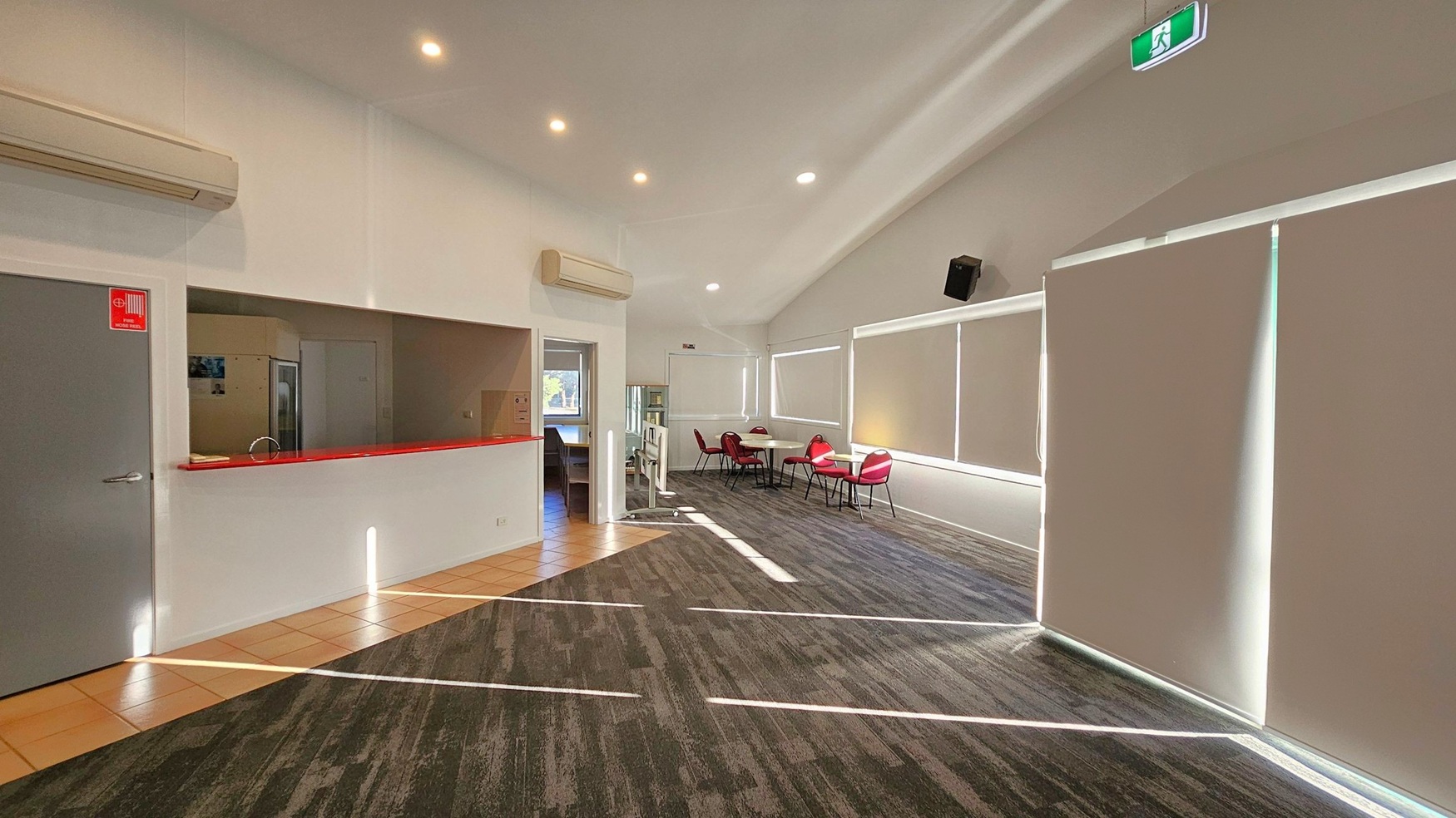 A spacious room with a high ceiling and recessed lighting. On the left, there's a grey door with a fire extinguisher sign and a service counter with a red edge. Above the counter is an air conditioning unit. The floor transitions from tiled to carpet. On the right, large windows are covered with blinds, and there are tables with red chairs. An emergency exit sign is visible near the ceiling.