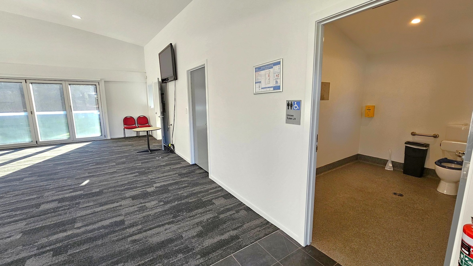A room with a grey carpeted floor and a white ceiling. On the right, an open door reveals a unisex accessible toilet with a toilet, grab bar, bin, and toilet brush. A sign outside the door indicates the toilet's accessibility. On the left, there are large windows with blinds, two red chairs, and a small table. A wall-mounted TV is above the chairs. The floor transitions from carpet to tiles near the toilet entrance.
