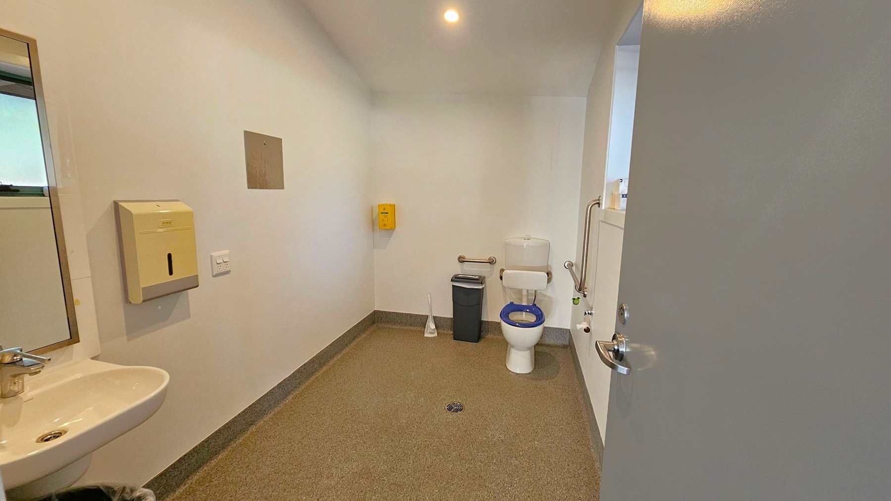 A bathroom with a grey speckled floor and white walls. On the left, a small sink with a silver tap is mounted on the wall, accompanied by a soap dispenser and a mirror. In the centre, a toilet with a blue seat is positioned against the back wall, with a grab rail beside it. A black bin and a toilet brush are nearby. On the right, the door is partially open, revealing a handle. The ceiling is plain with a single recessed light.