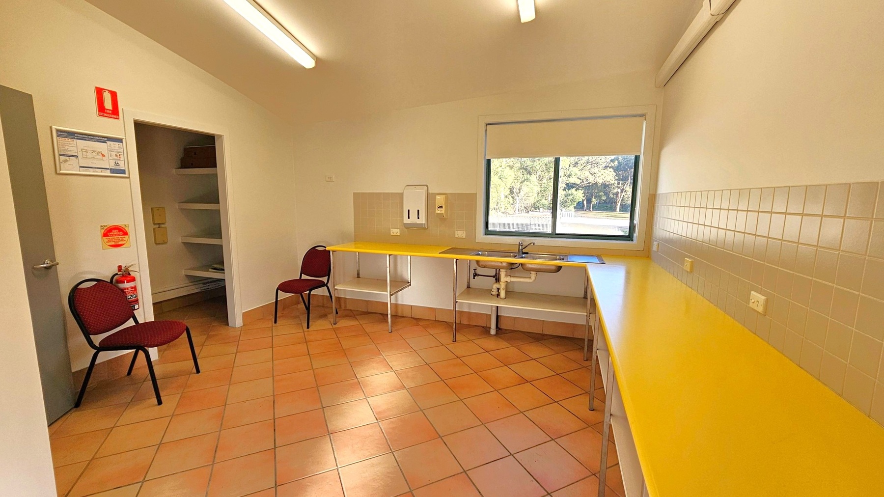 A room with terracotta floor tiles and a high ceiling with fluorescent lights. On the left, there are two brown chairs and a fire extinguisher near a door. A wall-mounted notice board and a fire safety sign are above. In the middle, a yellow countertop with a sink and a soap dispenser is beneath a window showing trees outside. The right side has a continuation of the yellow countertop with beige tiled backsplash and power outlets.