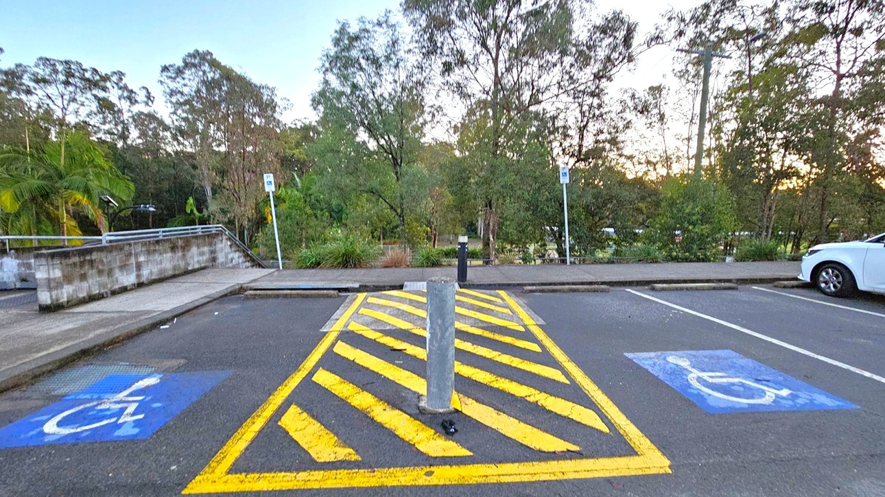 A parking area with two marked disabled parking spaces on either side, indicated by blue paint with a white wheelchair symbol. In the centre, there is a yellow-striped zone with a concrete bollard. A white car is parked on the right. The background features a footpath and dense greenery with various trees and plants. A low stone wall is visible on the left, and a signpost is present near the centre.