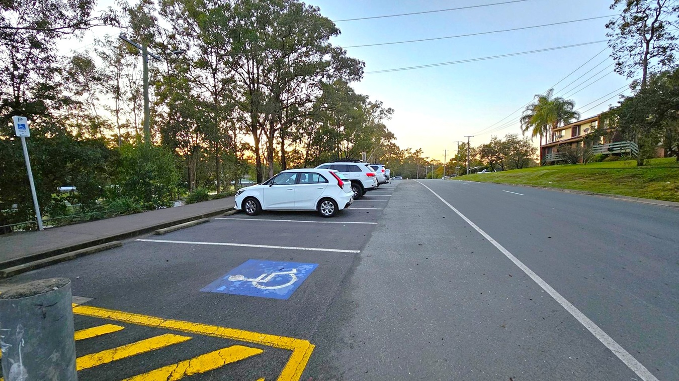 A row of parked cars is on the left, with a white car in the foreground occupying a disabled parking space marked with a blue symbol. A signpost indicates the parking restriction. Trees line the left side, providing a natural backdrop. On the right, a road stretches into the distance, bordered by a grassy area with a building partially visible. Power lines run overhead, and the pavement is clear and well-maintained.
