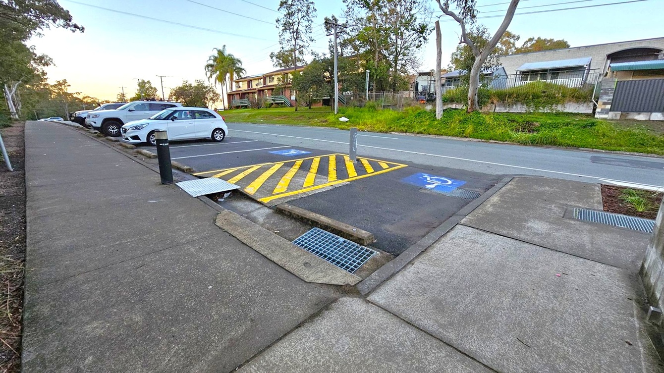 A concrete footpath runs along the left side, with a row of parked cars adjacent to it. The parking area includes a space marked with yellow diagonal lines and a disabled parking symbol. Across the road, there is a grassy verge with trees and a building in the background. The road is clear, and the area appears to be a suburban setting.
