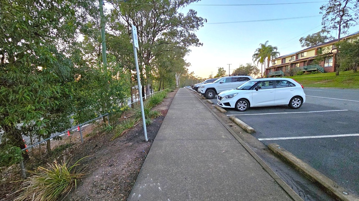 A concrete footpath runs through the centre, flanked by a row of parked cars on the right, including a white hatchback. On the left, there are trees and shrubs, with a metal fence partially visible. In the background, a two-storey building with a sloped roof and balconies is seen on the right, along with a few palm trees. The sky is clear, and the area is well-lit, suggesting early morning or late afternoon.