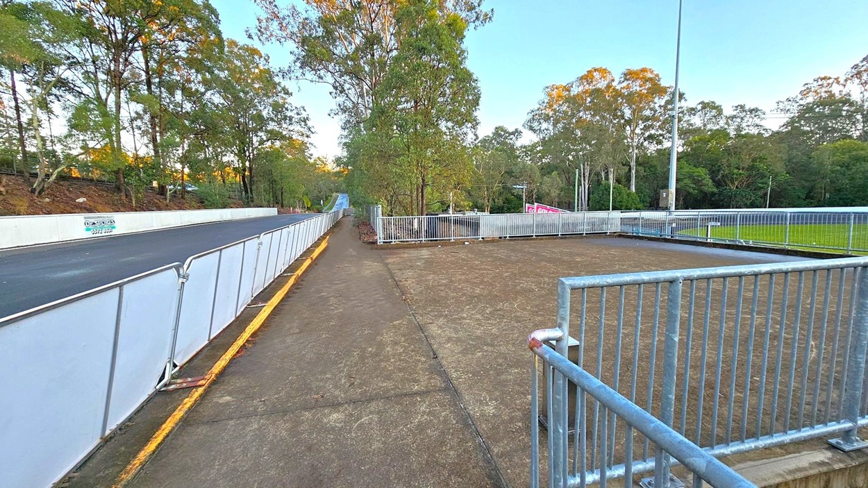 A paved road on the left is lined with white barriers and bordered by a row of trees. On the right, a fenced-off dirt area is visible, with a tall lamp post and more trees in the background. A concrete path runs alongside the road, with scattered leaves on the ground. The scene is surrounded by dense greenery, and a sign with text is partially visible in the distance.
