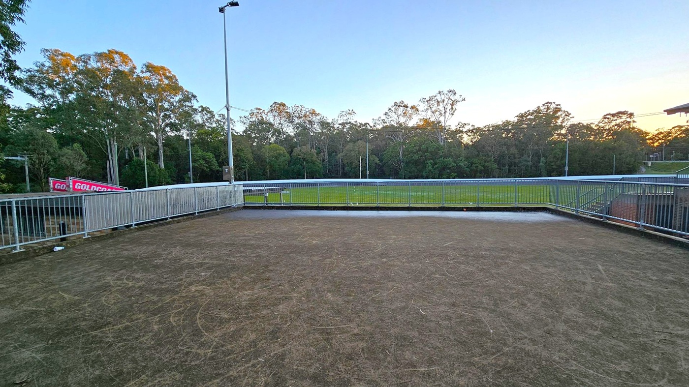 A fenced area with a rough, dark surface in the foreground, possibly a sports or viewing platform. Beyond the fence, a grassy field stretches out, bordered by a line of tall trees. A tall light pole stands to the left, and a sign with "GOLDCOAST" is partially visible. The sky is clear, with a hint of sunset on the horizon.