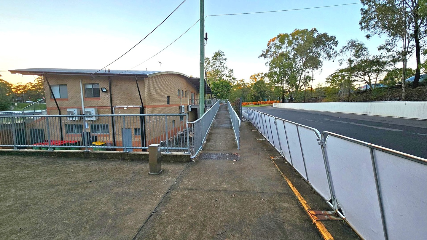 A pathway with metal railings runs through the centre, flanked by a brick building on the left and a road with temporary barriers on the right. The building has a flat roof and several windows, with air conditioning units visible. Trees are in the background, and the ground is covered with a mix of concrete and patches of grass. Overhead, power lines are strung between poles. The scene is lit by the soft light of either dawn or dusk.