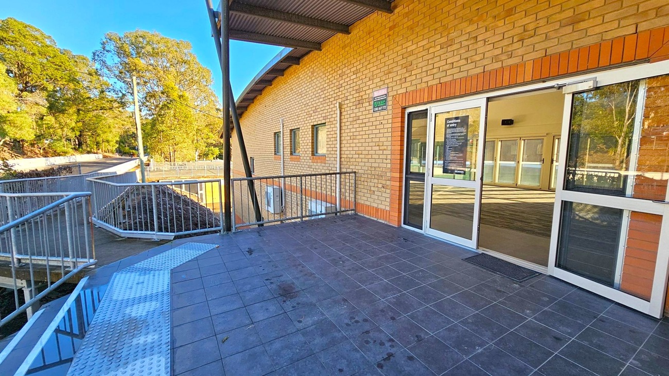 A brick building with a sloped roof on the right, featuring a glass door with a sign. The floor is tiled in dark grey. A metal railing lines the edge of the walkway, leading to a ramp on the left. In the background, there are tall trees and a clear blue sky. A small puddle and a single leaf are visible on the ramp.