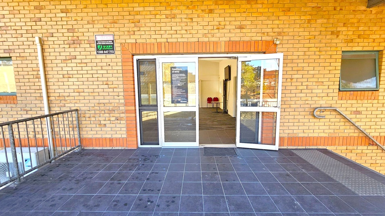 A brick building with a double glass door entrance in the centre, partially open. Above the door, a sign with security information is visible. Inside, a small table and two red chairs are seen. The floor outside is tiled in dark grey, with a metal ramp on the right. A metal railing is on the left, and a small window is on the right side of the wall.