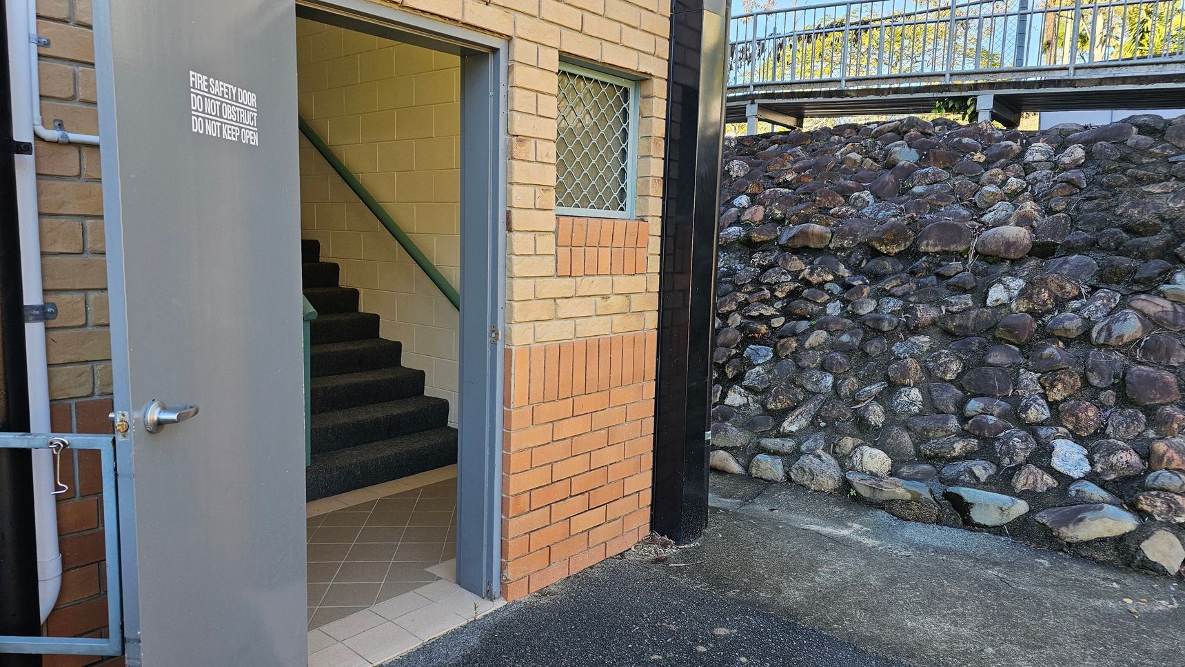 A grey fire safety door is open, revealing a staircase with dark carpet and a green handrail inside a brick building. The wall has a small window with a metal grid. To the right, a stone retaining wall is visible, with a metal railing above it. The ground is a mix of concrete and asphalt.