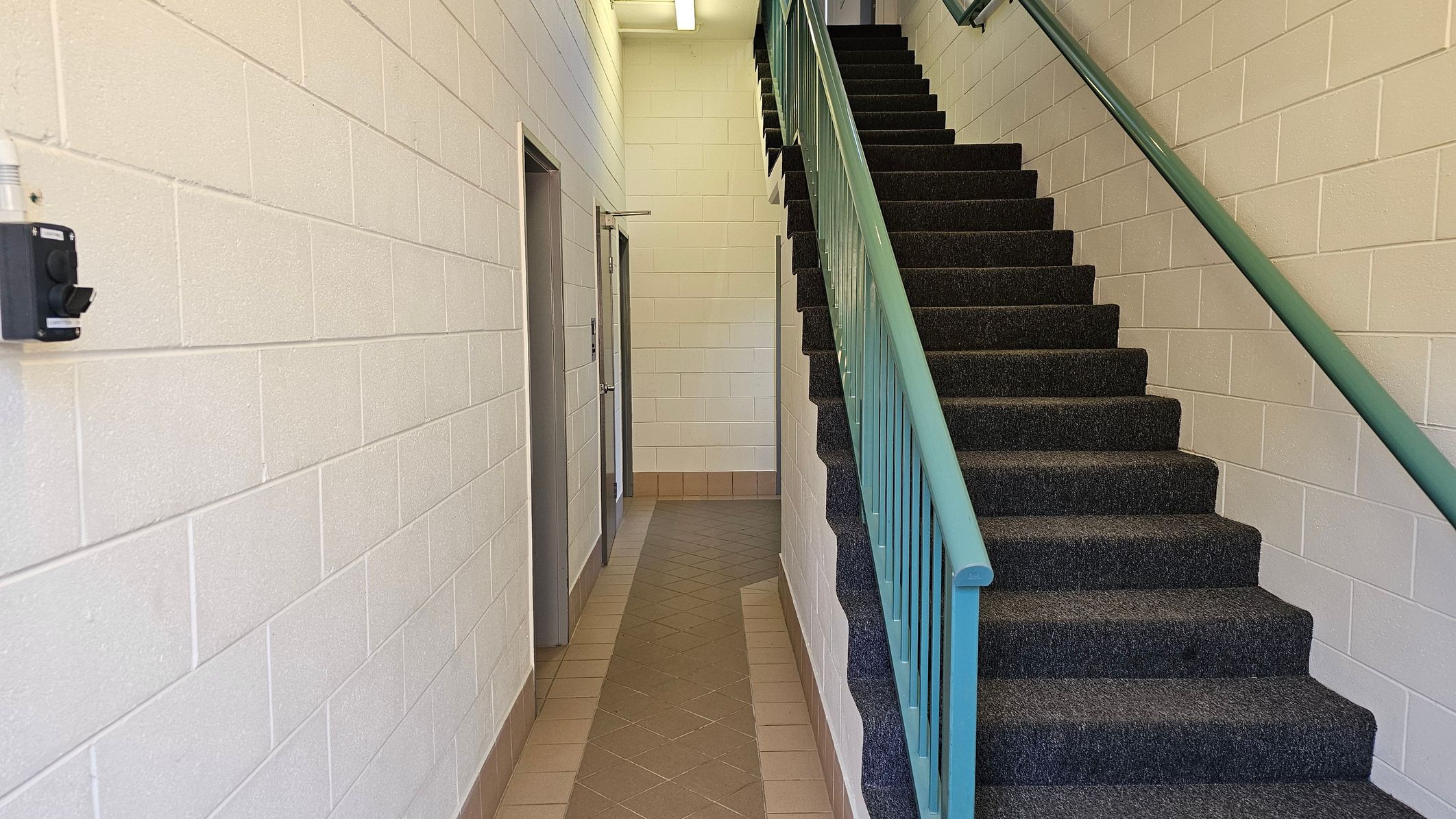 A stairwell with dark carpeted steps and a green handrail on the right. The walls are made of light-coloured bricks. On the left, there is a corridor with a tiled floor leading to a door. A black device is mounted on the left wall. The ceiling has a fluorescent light fixture.