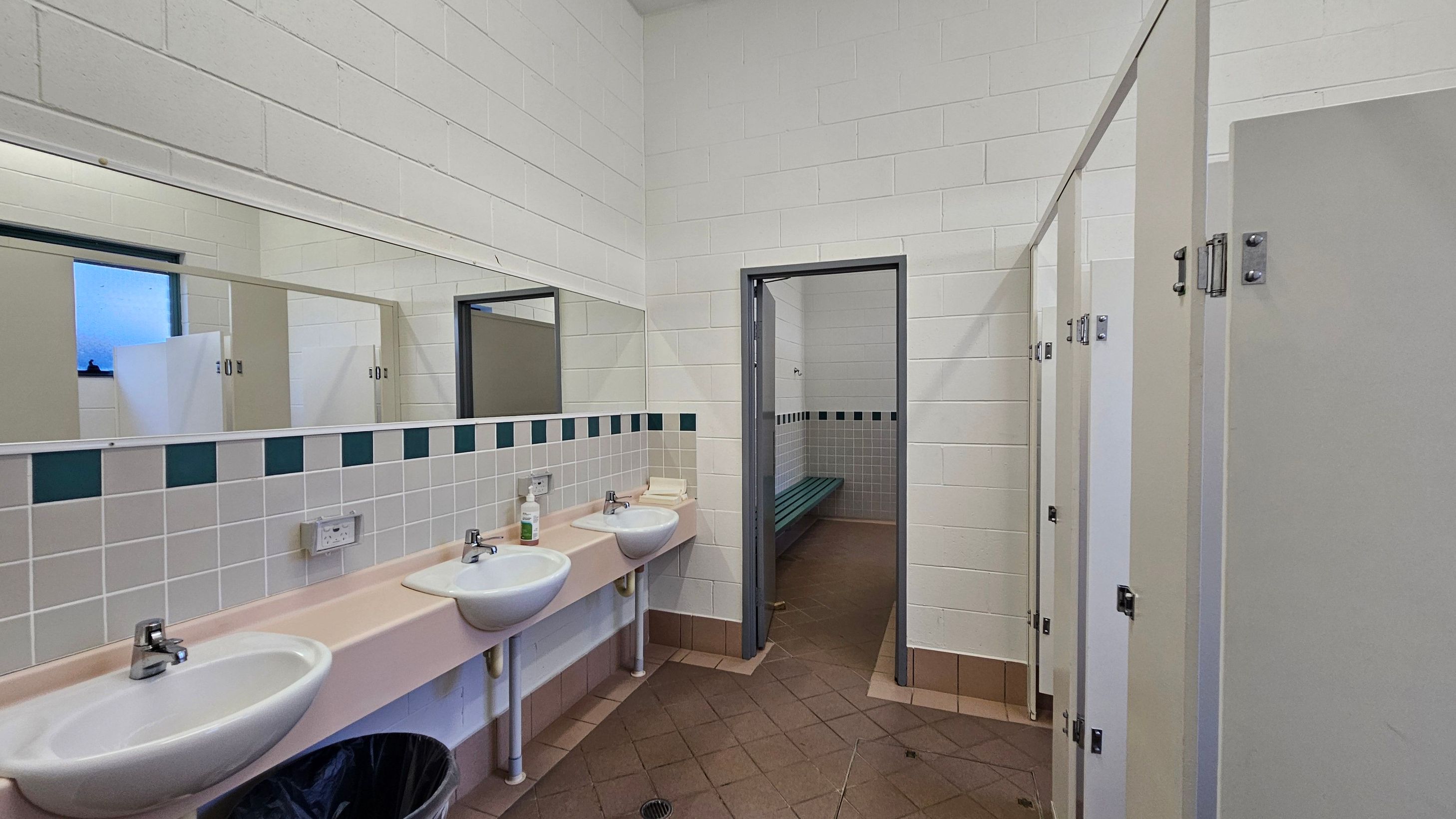 A public restroom with white tiled walls and a beige tiled floor. On the left, there are two white sinks with a pink countertop, a mirror above, and a soap dispenser. A black bin is beneath the sinks. In the middle, an open doorway leads to a room with a green bench and more tiles. On the right, there are several closed stall doors with metal locks. The ceiling is white and flat.