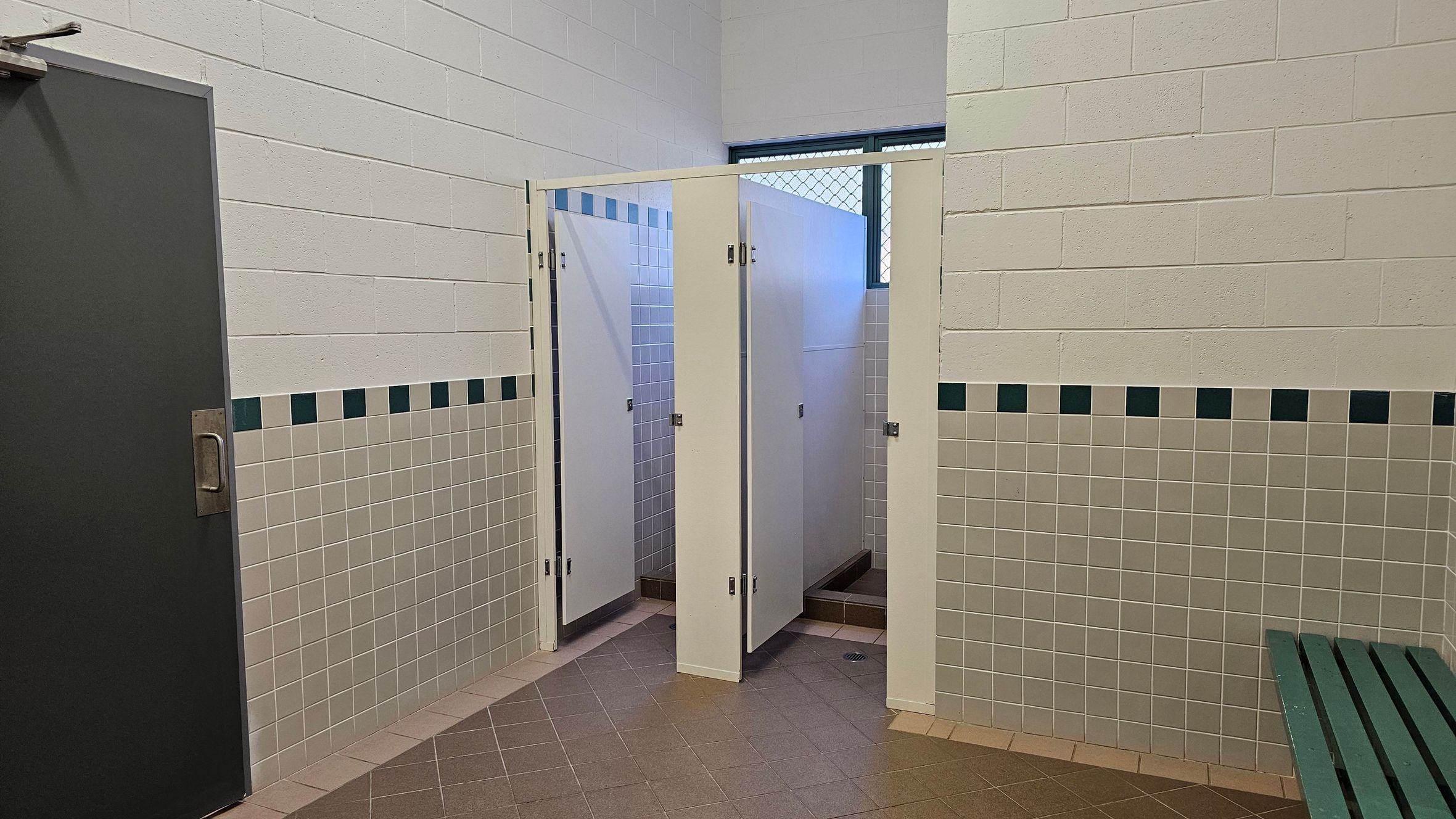 A public restroom with a grey door on the left, featuring a metal handle. In the centre, there are three shower stalls with white doors, slightly ajar, revealing tiled interiors. The walls are tiled with beige and green square patterns. A small window with a metal grid is above the stalls. On the right, a green bench is visible. The floor is covered with brown tiles arranged diagonally.