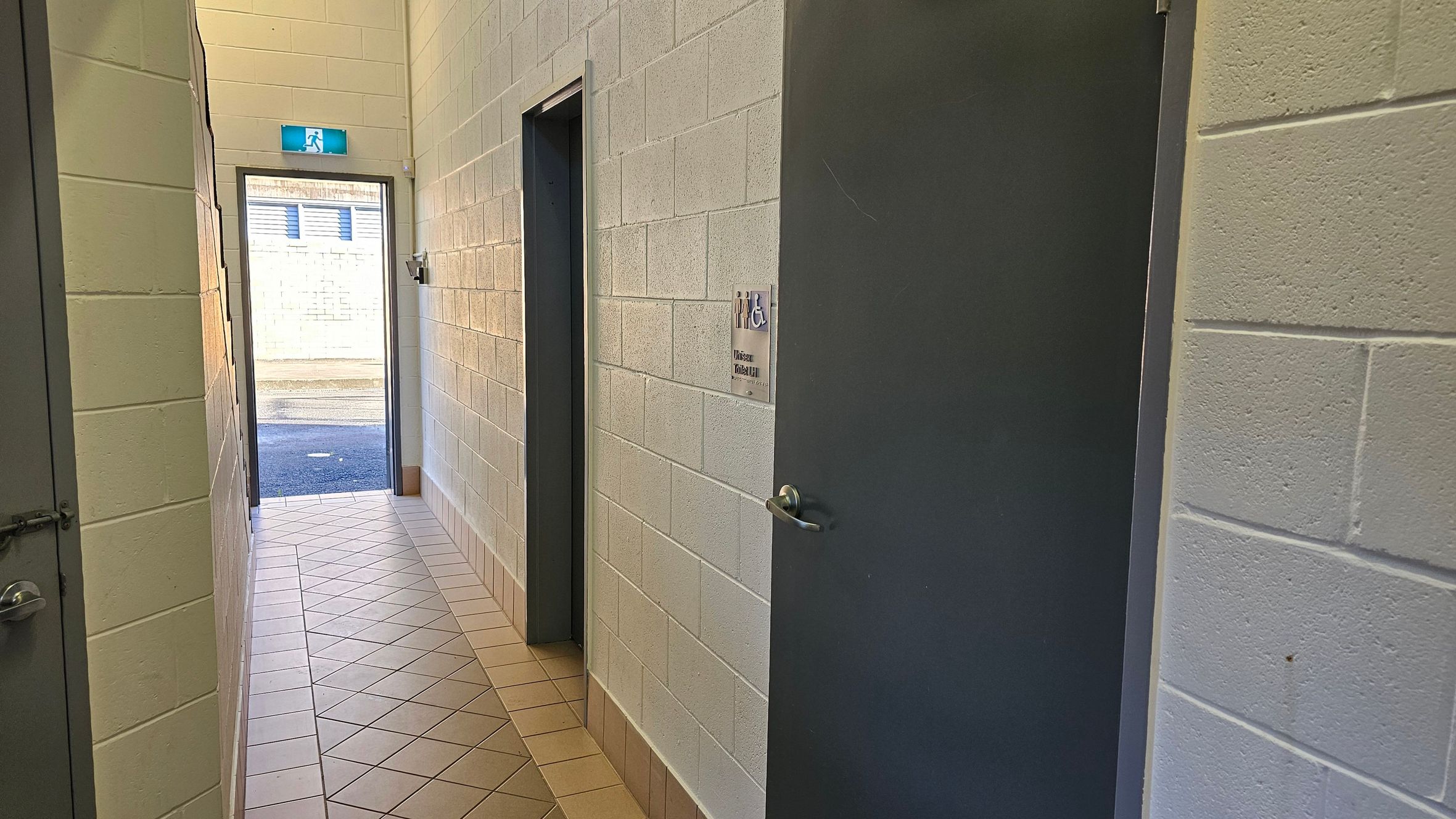 A narrow corridor with light-coloured brick walls and a tiled floor. On the right, there is a dark door with a sign indicating a unisex accessible toilet. Further down the corridor, another doorway is visible. At the end of the corridor, an open door leads outside, where a brick wall and a small section of pavement can be seen. An emergency exit sign is above the door.