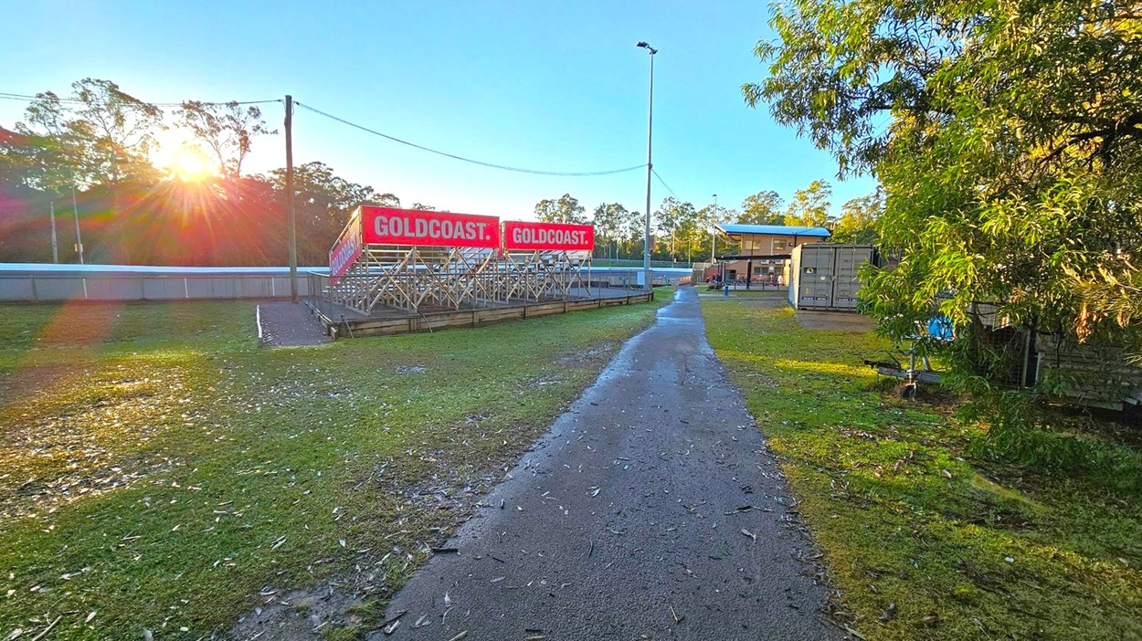 A paved path runs through the centre, flanked by grass. On the left, there are metal bleachers with red signs reading "GOLDCOAST." The sun is setting behind trees, casting long shadows. On the right, a building with a blue roof and some shipping containers are visible. A tall streetlight stands near the centre. Trees and foliage are scattered throughout the background.